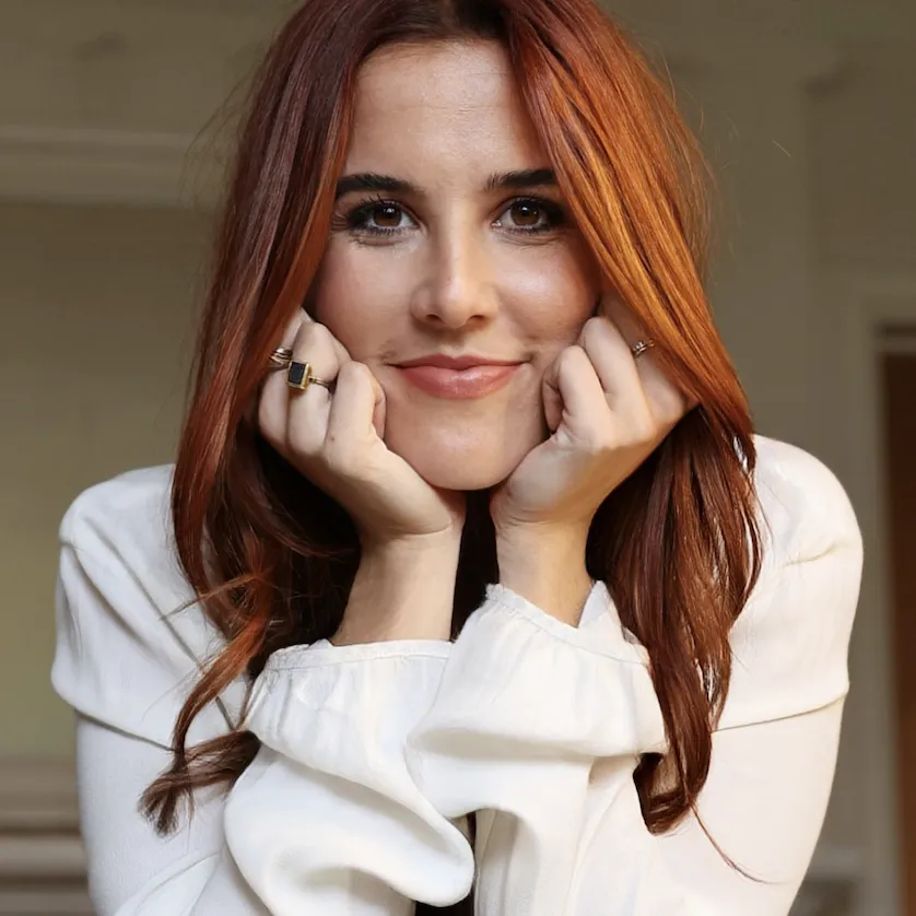 Smiling woman with long red hair resting her chin on her hands, wearing a white top.